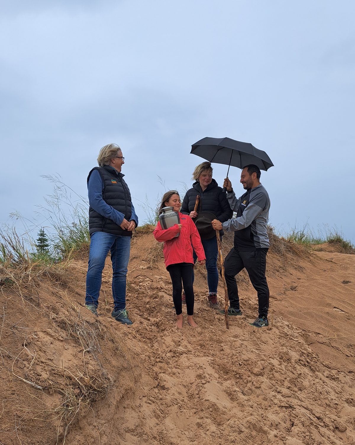 A family on a red sand dune with an urn, sharing an umbrella on a cloudy day